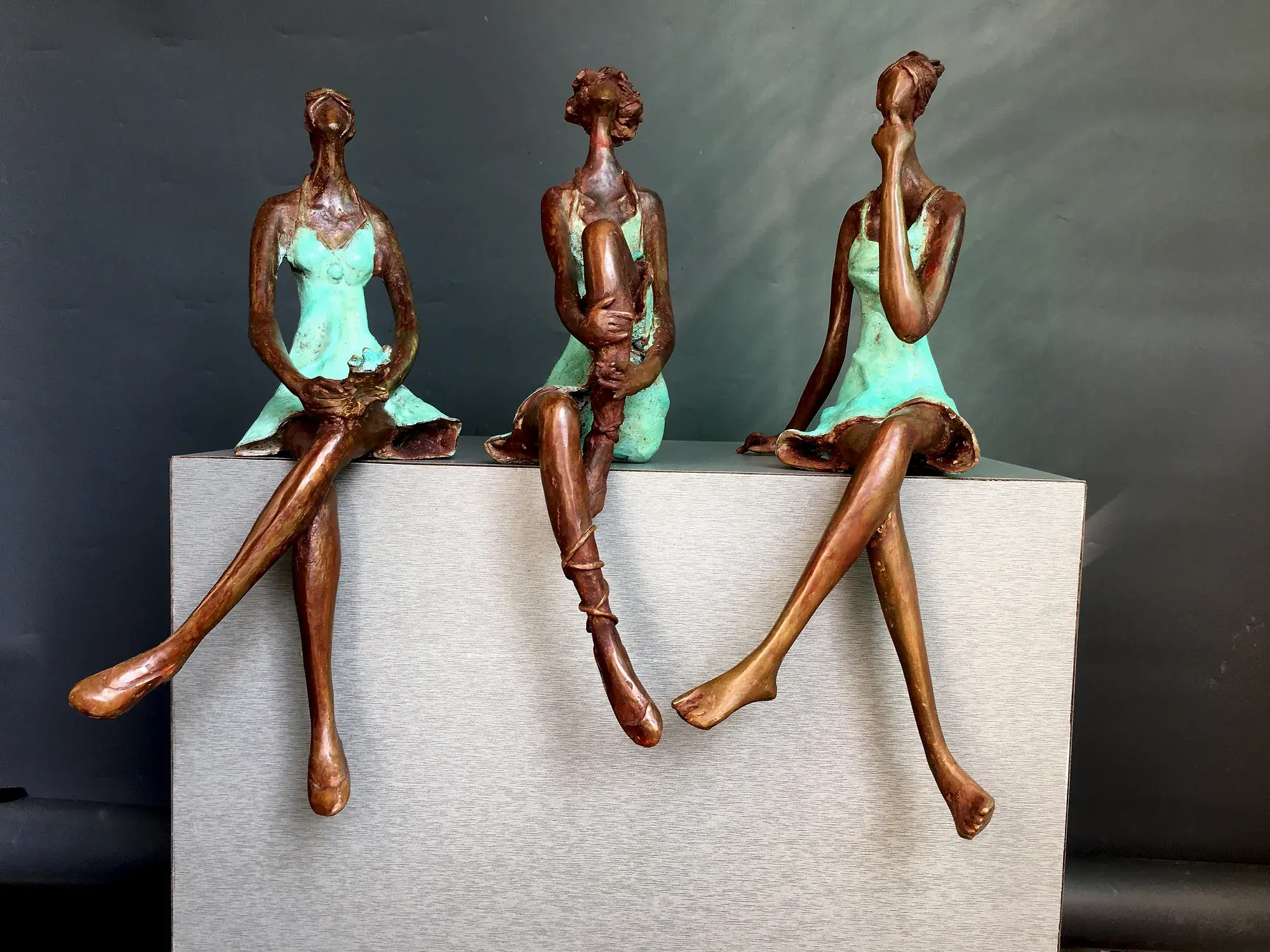 Conversation III — three small bronze figures in mint-green dresses seated on a plinth edge, legs dangling