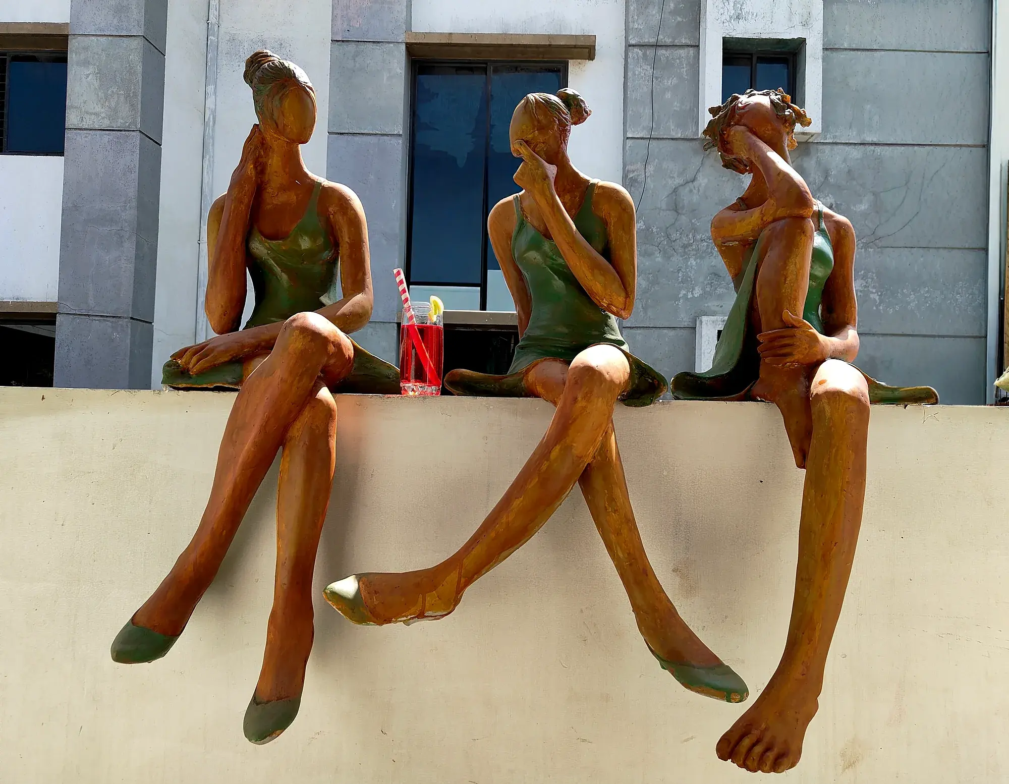 Conversation — three life-size figures in green swimsuits seated on an outdoor ledge, one holding a drink, building backdrop