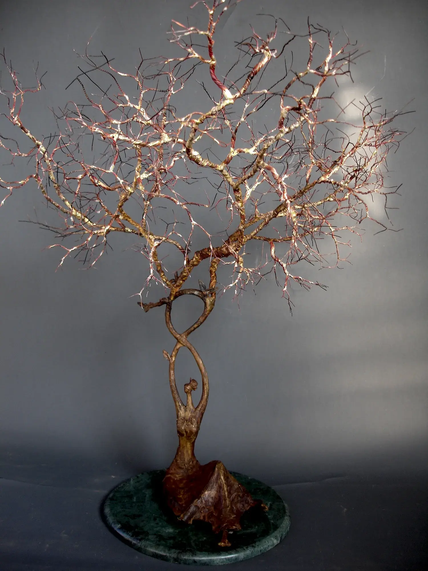 Kalpavruksha — bronze and brass-wire tree sculpture rooted in a woman's silhouette, bare branches spreading above a round dark-green marble base