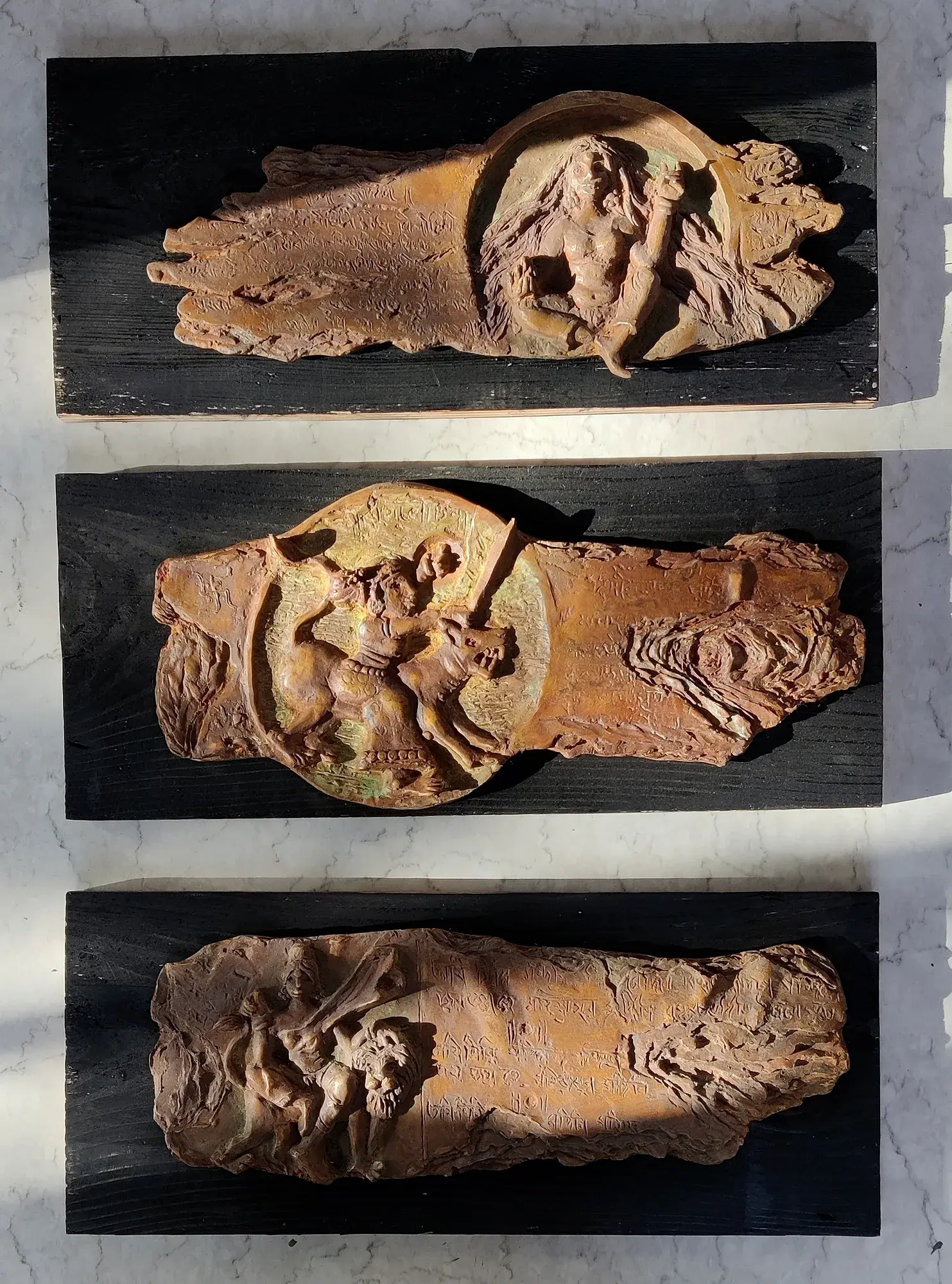 Manuscript Series — three terracotta and bronze relief panels on dark wood, each with a circular relief scene of a goddess slaying a demon