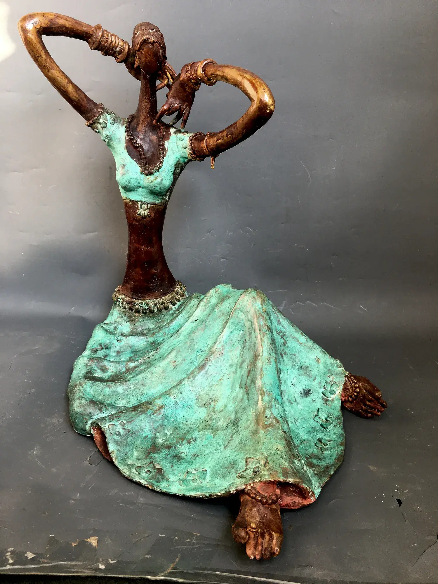 Bronze seated figure in mint-green patina dress, arms raised in an overhead loop, adjusting a necklace