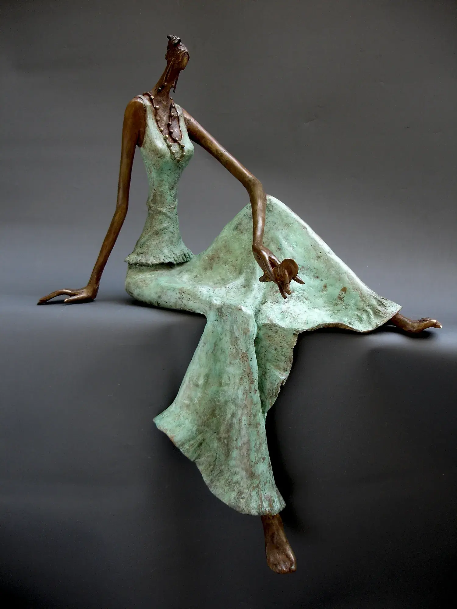 Bronze seated figure in mint-green patina dress, one arm arced overhead, the other hand holding a flower to the chest